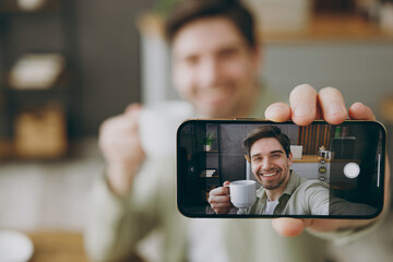 Clsoe up young fun man wears casual clothes doing selfie shot on mobile cell phone post photo on social network drink tea sits alone at table in coffee shop cafe restaurant indoors rest relax inside.