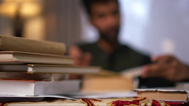 Close-up books pile and blurred male adult student opening book reading sitting at background. Intelligent unrecognizable Middle Eastern man studying at home indoors