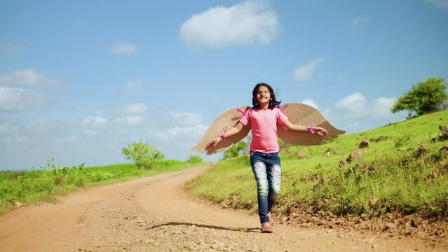 Happy smiling girl kid with artificial wings by running by dancing near green mountain - concept of freedom, imagination and enjoyment