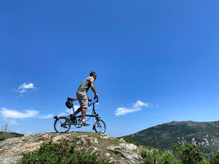 Fototapeta na wymiar Un Uomo sulla sua bici pedala sopra una roccia, isolata su sfondo cielo azzurro