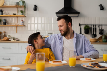 A caring father sits with his son at the kitchen table. A little boy is sitting in the kitchen with his dad eating sandwiches.