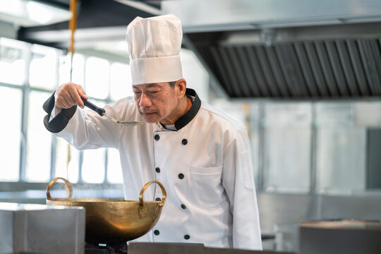 Professional senior Asian chef man in dress white uniform tasting delicious food in kitchen restaurant. Cooking and food concept