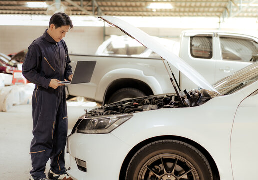 Auto mechanic working at automobile repair garage examining engine using notebook computer program. Asian repairman inspecting broken car for fixing or maintenance at automotive repair service shop.
