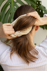Naklejka premium Blonde woman making a ponytail with beige scrunchie, shot from behind, focus on hands, photo in movement. Monstera's leaf in the background.
