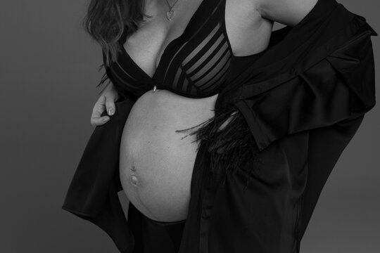 Cheerful young pregnant female touching belly standing against grey wall, pregnant woman, black and white photo