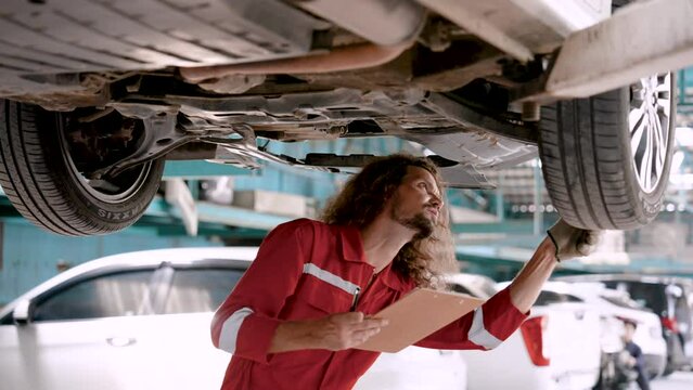 Caucasian man in dust suit from car repair A mechanic holding a checkbook examines the undercarriage of a raised car to facilitate repair. Use gloved hands to check the condition of the wheels.
