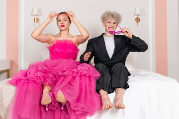 A Glamour Girl and a man looking like a doll, sitting on a bed. Doll couple dressed in long pink...