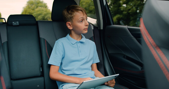 Good-looking happy smiling 10-year-old boy browsing apps on his tablet gadget and watching at the nature through car window while driving on the car's backseat - Powered by Adobe