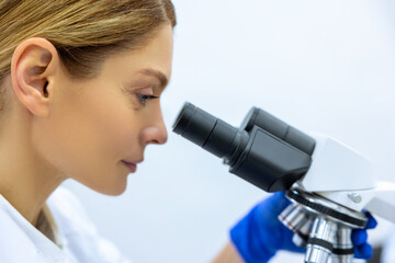 Blonde woman lab assistant in blue gloves looks in a microscope, chemical formula in lab test room