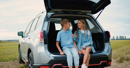 Lovely cheerful amicable 10-12-year-old brother and sister sitting in auto trunk surrounding green lawn and enjoying communication between themselves