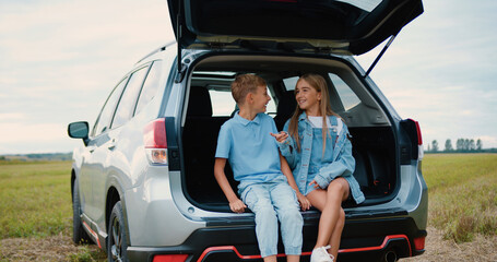 Lovely cheerful amicable 10-12-year-old brother and sister sitting in auto trunk surrounding green...