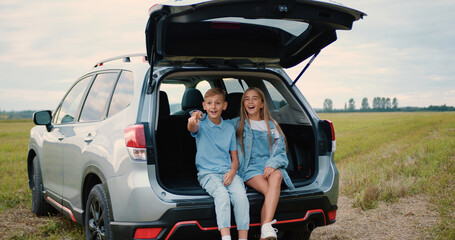 Road trip concept where good-looking positive friendly two children sitting in car trunk among...