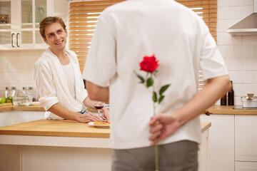 young gay couple looking at boyfriend and holding rose behind his back for surprise in the kitchen