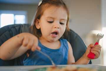 Cute adorable baby girl enjoying breakfast eating with fork from a plate at home kitchen. People lifestyle concept. Happy lifestyle. Baby feeding concept. Food