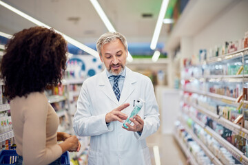 A male pharmacist holding a new product for skin care and showing it to a female customer.