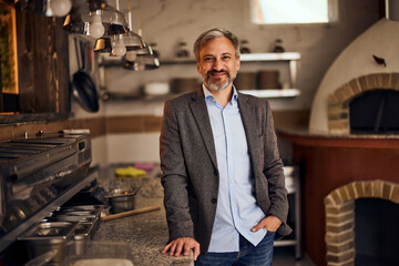 A senior male pizzeria owner standing in front of the pizza oven, smiling at the camera. - obrazy, fototapety, plakaty