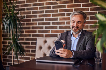 A smiling senior male restaurant owner using a mobile phone in front of a brick wall.