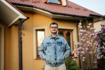 Portrait of stylish man in glasses and jeans jacket posed against his house.