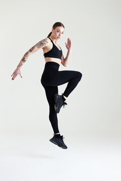 A young girl with brown hair, with a tattoo in a black top and leggings on a white background. Sports, fitness, jumping