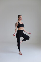 A young girl with brown hair, with a tattoo in a black top and leggings on a white background. Sport, fitness, standing with folded arms. Yoga