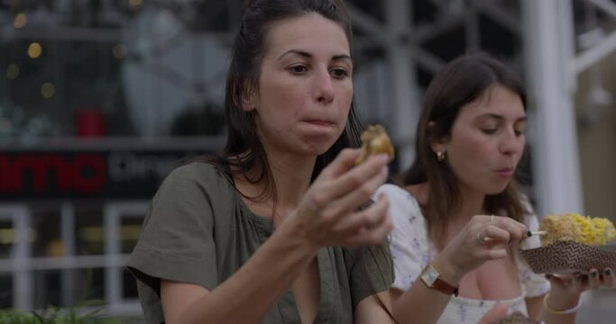 Two young mothers eat street food outdoors - share with kids - close up on face