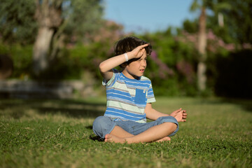 Young boy sitting cross-legged on the grass, shielding his eyes from the sun with his hand as he gazes into the distance. How exposure to natural light can impact children's vision and overall health