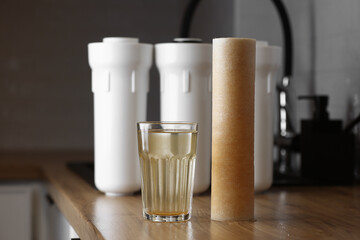 Water filter cartridge used and a glass of rusty water brown coloring on wooden table at kitchen interior. Plastic set with three filter cartridges reverse osmosis water purification system.