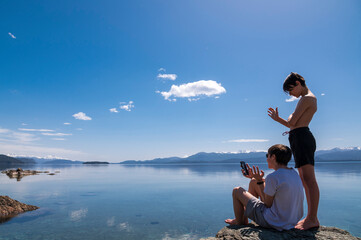 Fototapeta premium Dos hermanos hablando por video chat con el celular desde la inmensidad de la naturaleza en medio del lago y las montañas. 