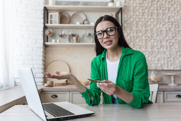 Young woman worried about credit card problems. He sits at home in the kitchen with a laptop, holds a card in his hands, spreads his hands and looks sadly at the camera.