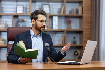 A young man pastor of a church, community sits in the office with a laptop and a microphone and...