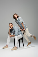 cheerful teenager leaning on chair next to happy father sitting and smiling on grey.