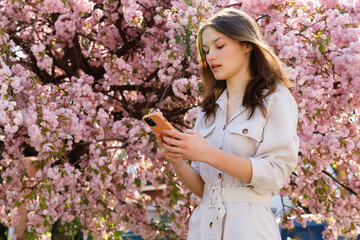 Adorable Young Lady with Modern Smartphone in Hands Smiling on Fresh Air. Portrait of Positive Caucasian Brunette Girl Posing Near Blooming Sakura Tree