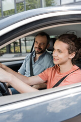 happy teenage boy holding steering wheel while driving car next to dad.