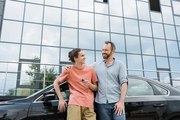 Gordijnen happy bearded man putting hand on shoulder of cheerful teenage son holding car key near automobile. © LIGHTFIELD STUDIOS