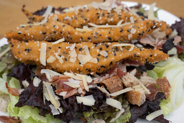 Food background. Caesar salad. Top view of fresh a salad with lettuce, parmesan cheese, bread croutons, fried chicken fingers breaded with sesame seeds and crispy bacon.