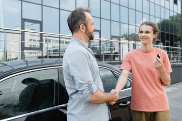 Fototapeta na wymiar happy bearded man shaking hands with cheerful teenage son holding car key near automobile.