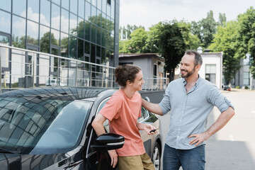 happy bearded father looking at cheerful teenage son holding car key near automobile.