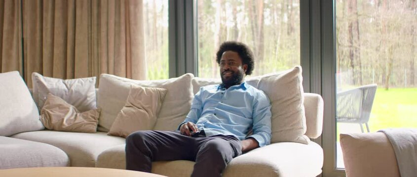 Portrait of Black African-American male spending his free time alone at home, sits on sofa, turns on TV