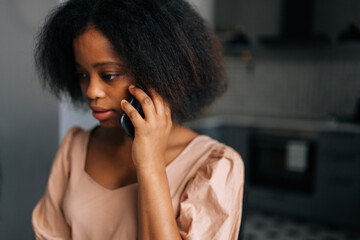 Closeup face of curly young African American woman talking on mobile phone at home. Close up portrait of attractive black female calling smartphone indoor. People and technology concept.