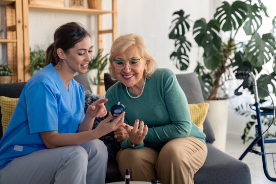 Young supportive female nurse teaching elderly senior woman to use blood sugar device at home. General practitioner with homecare patient. Home care, help, support and empathy for older adults.