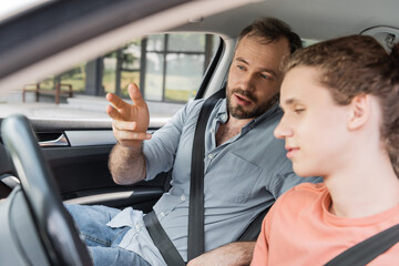 bearded dad gesturing while explaining to teenage son how to drive car.
