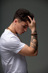 Portrait of a young brunette boy in a white t-shirt with a snake tattoo on his arm, beads on his neck, a bracelet on his arm, fashionable short haircut. Contemporary urban fashion. Gray background.