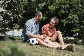happy teenage boy sitting near soccer ball with father in green summer park.