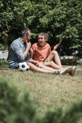 amazed teenage boy sitting near soccer ball with father in green summer park.