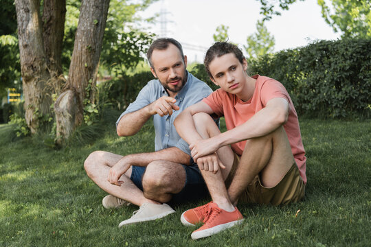 bearded man pointing away near teenager son while sitting together on green lawn.