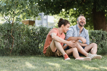 happy father laughing while sitting near teenage son on green lawn in park.