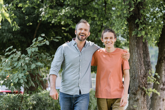 cheerful father with beard hugging overjoyed teenager son while walking in green park.