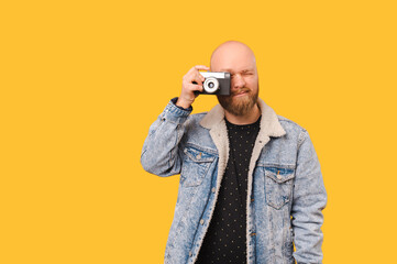 Fototapeta premium Young bald man wearing jeans jacket is taking a picture with a vintage camera over yellow background.
