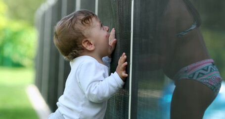 Obraz premium Cute baby standing by swimming pool fence protection. Toddler interacting with child