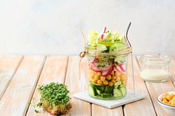 Healthy salad of cucumber, boiled chickpeas, radish, iceberg lettuce and microgreens in a glass jar on a wooden background. Healthy food, diet, vegan concept.